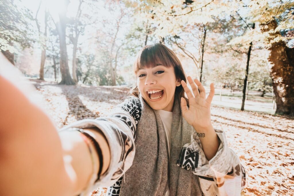 Una mujer sonriendo mientras se toma un selfie en un parque otoñal. Lleva bufanda y suéter, con hojas caídas y luz solar filtrándose entre los árboles.