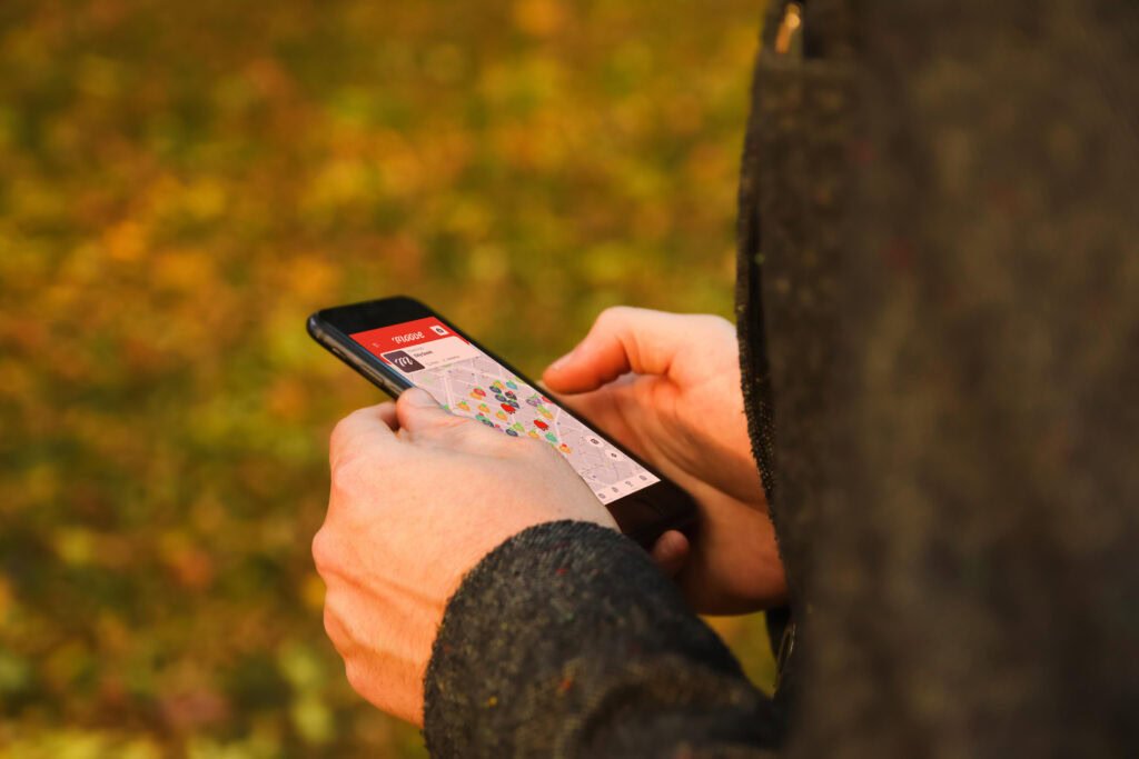 Primer plano de una persona sosteniendo un smartphone al aire libre, mostrando la aplicación Mooveteam con un mapa de geolocalización en la pantalla. De fondo, hojas de otoño creando una atmósfera cálida y otoñal.
