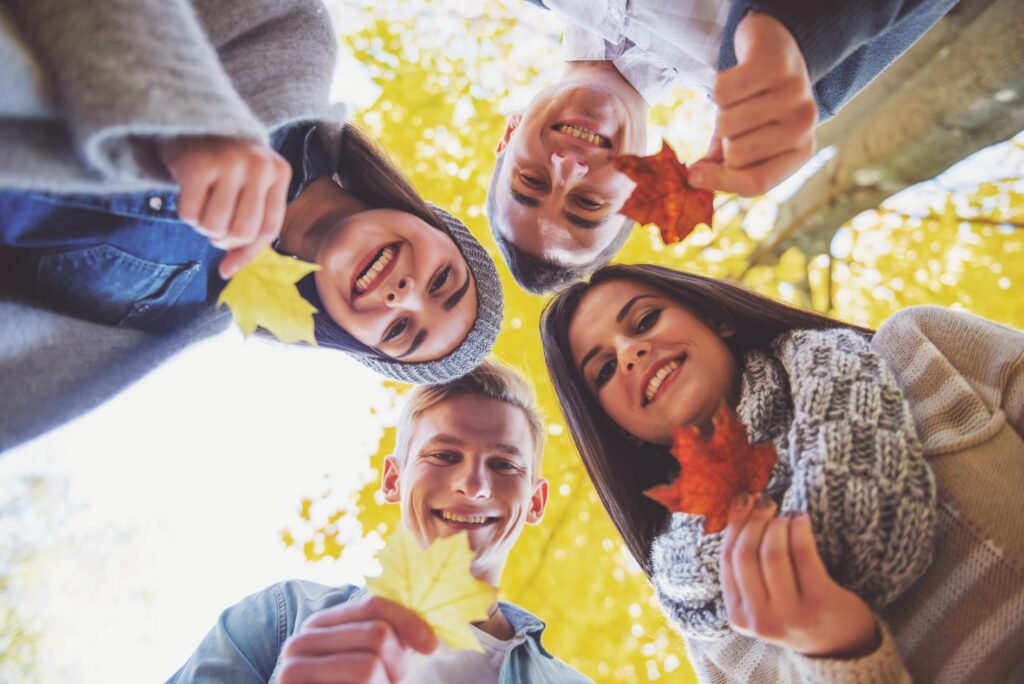 Un grupo de jóvenes sonrientes sosteniendo hojas de otoño en un parque. De fondo, follaje otoñal en tonos amarillos y naranjas.