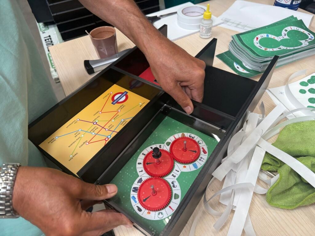 Close-up of a person assembling a Seconds2Survive EscapeBox, revealing internal puzzle components including red decoder dials and a map resembling a subway system. Surrounding the box are game cards, tape strips, and assembly tools on a work table.