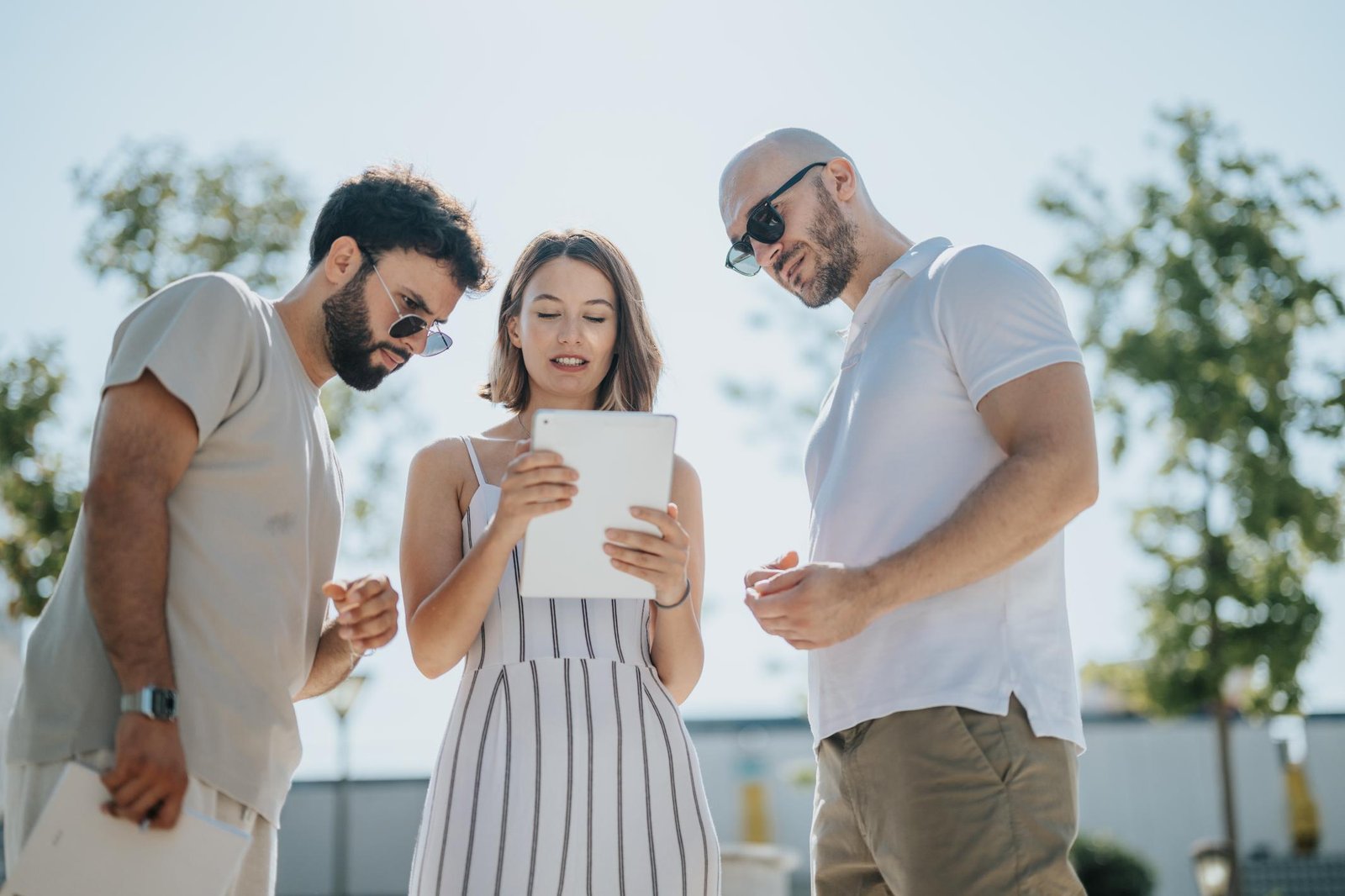 Three people participating in an outdoor team-building activity using a tablet. The sunlit setting suggests a casual, collaborative atmosphere as two men and a woman focus on the screen, potentially solving a puzzle or navigating a scavenger hunt challenge.