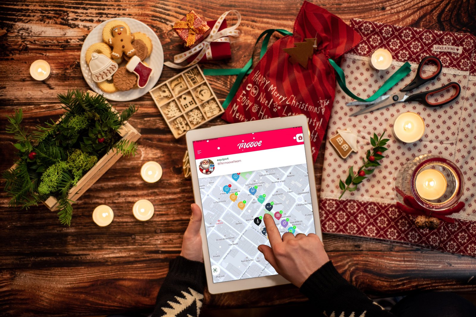A person wearing a festive sweater is using a tablet displaying a Mooveteam app with a city map and various interactive points marked. The tablet is surrounded by Christmas-themed decorations, including a red gift bag, holiday cookies, candles, scissors, and wrapped presents on a wooden table.
