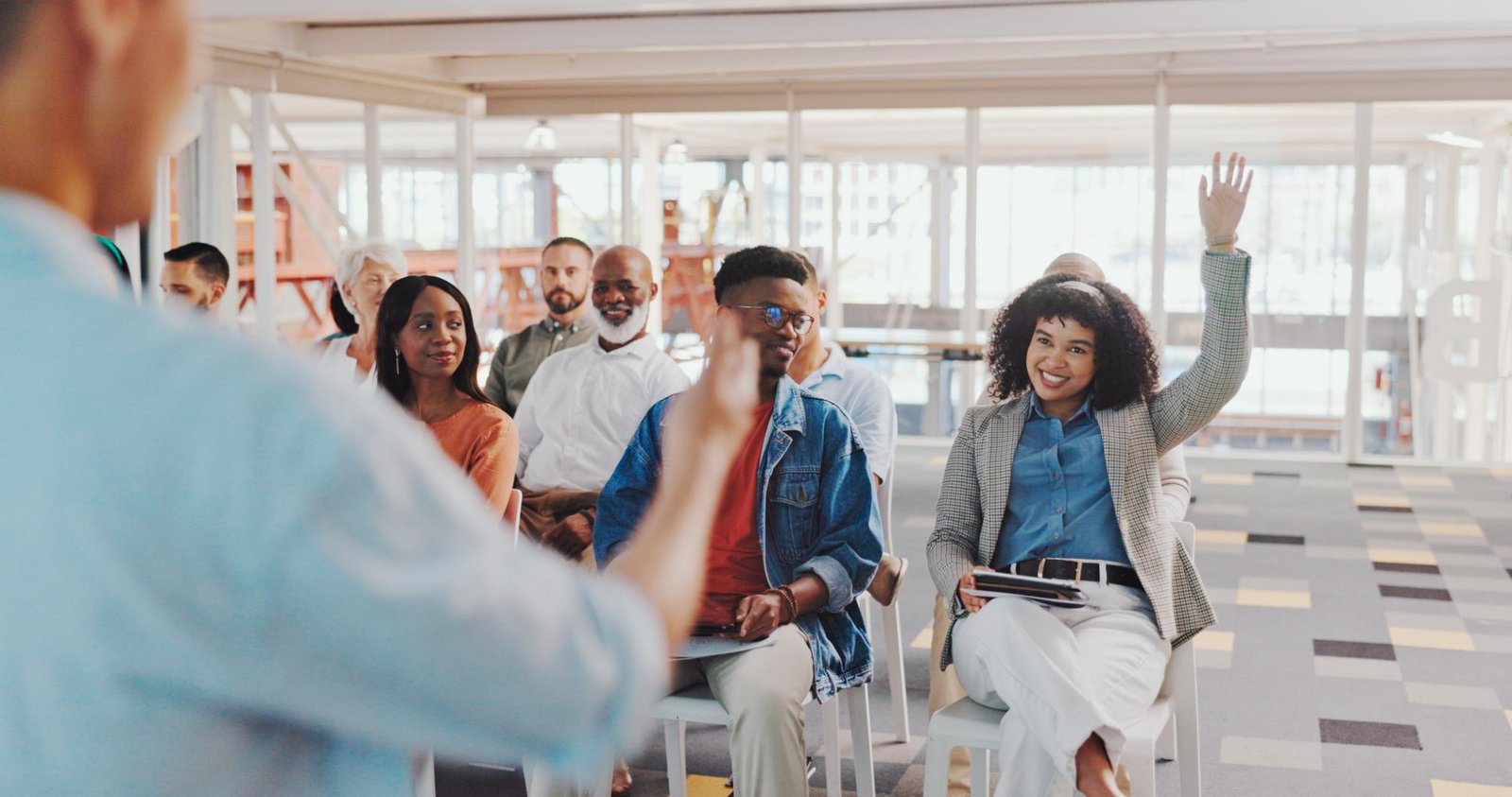 A diverse group of professionals actively participating in a corporate training session, raising their hands and engaging with the presenter.