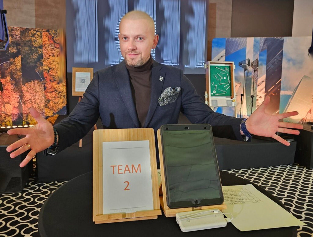 Event facilitator at Aparthotel Termy Uniejów presenting the 'Moove Your Mind' team-building challenge setup, featuring task stations with tablets and team labels to guide participants through interactive puzzles and QR code challenges."
