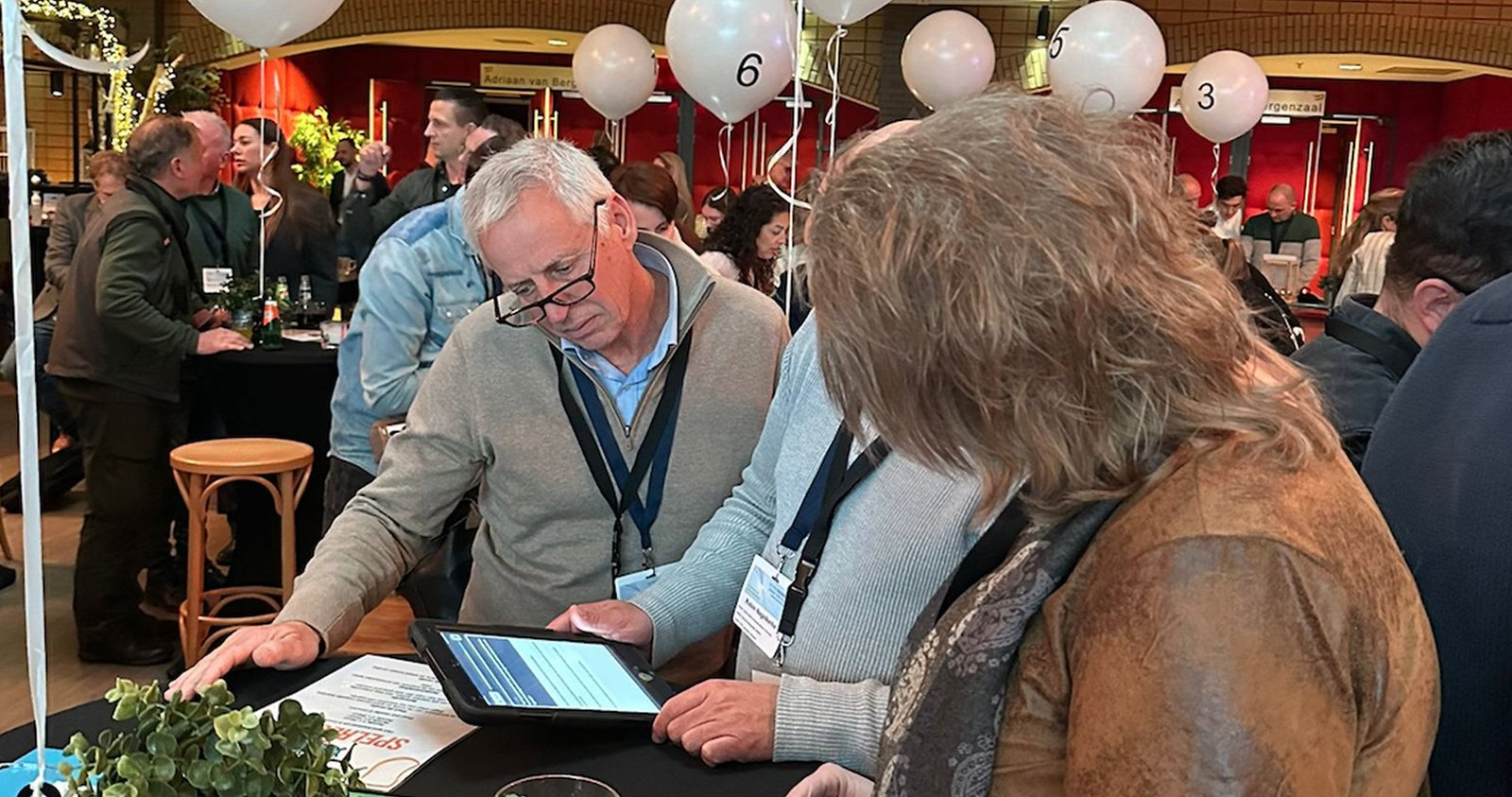 A group of participants engaging in the Turning Tables networking game at a corporate event. They are using a tablet to complete interactive challenges, with numbered balloons indicating different tables. The event is lively, with multiple discussions taking place in the background.