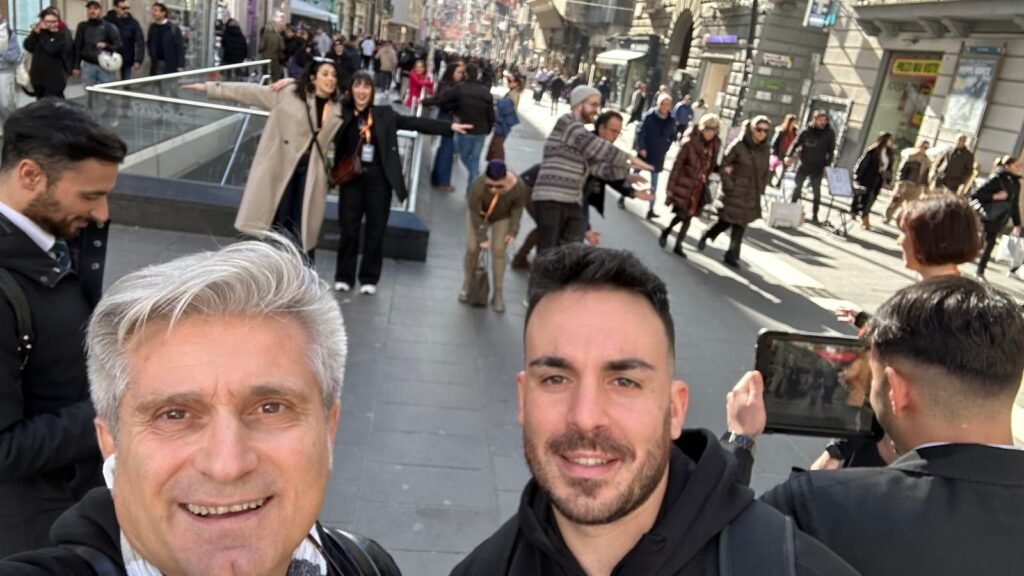 Team-building activity by EAmilano on a lively street in Naples, Italy. Participants are smiling, taking part in a gamified urban challenge, with some using a tablet and others posing energetically in the bustling pedestrian area. The backdrop shows local shops, historic buildings, and passersby enjoying the day.