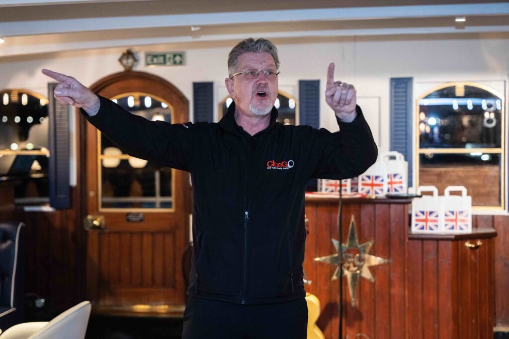 A ClueGO team leader giving instructions with dynamic gestures during a team-building briefing indoors. Union Jack-themed welcome packs are visible in the background.