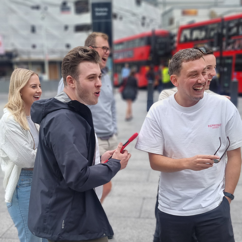 Group of young adults laughing and interacting with a mobile device during a ClueGO team-building game on a London street, with red double-decker buses in the background.